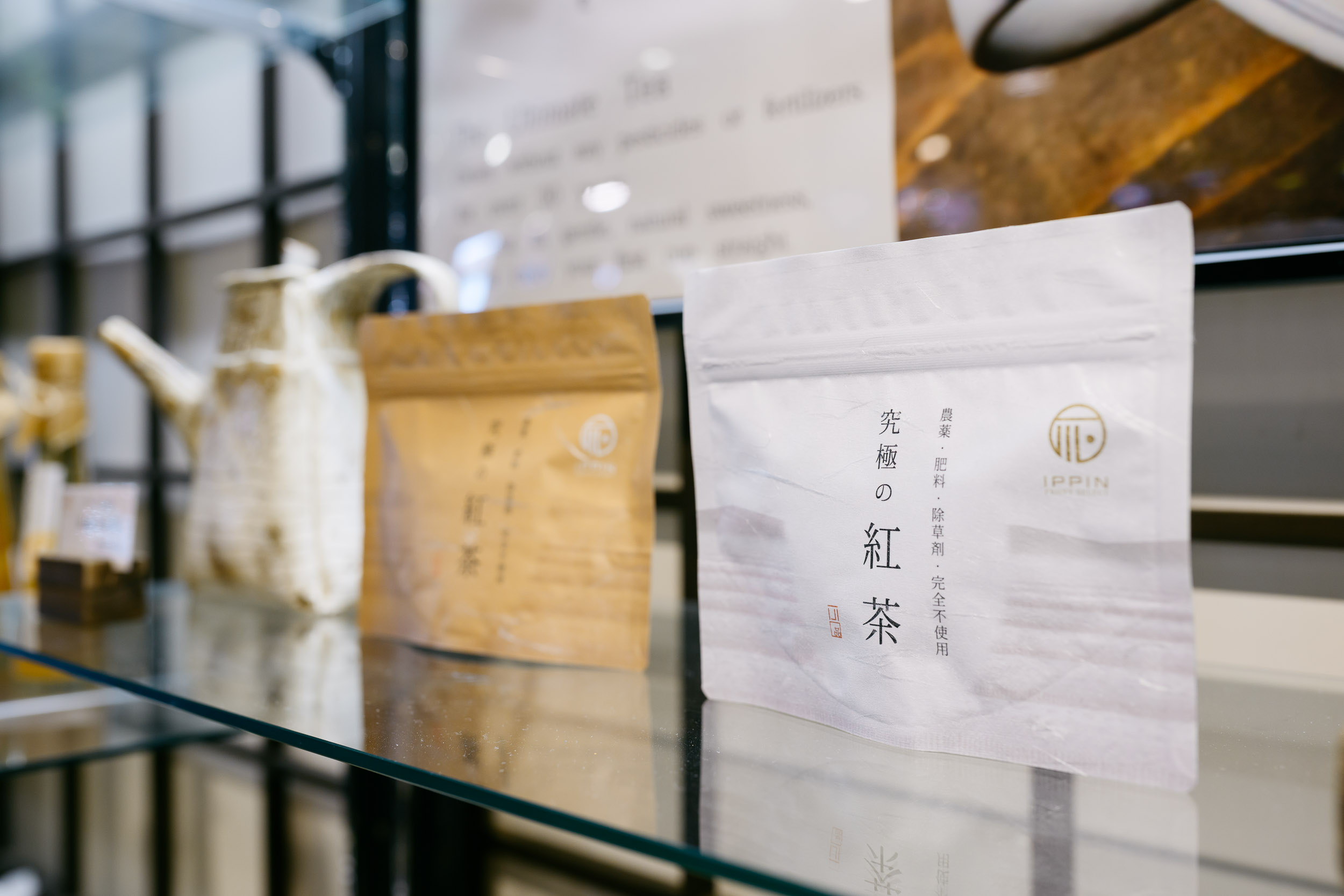 下田時計台フロントのガラス棚に並ぶ「IPPIN PROJECT」の「究極の紅茶」パッケージ。手前には白、奥には茶色のチャック付きスタンド袋が並び、背景には急須や店内のディスプレイが写っている。