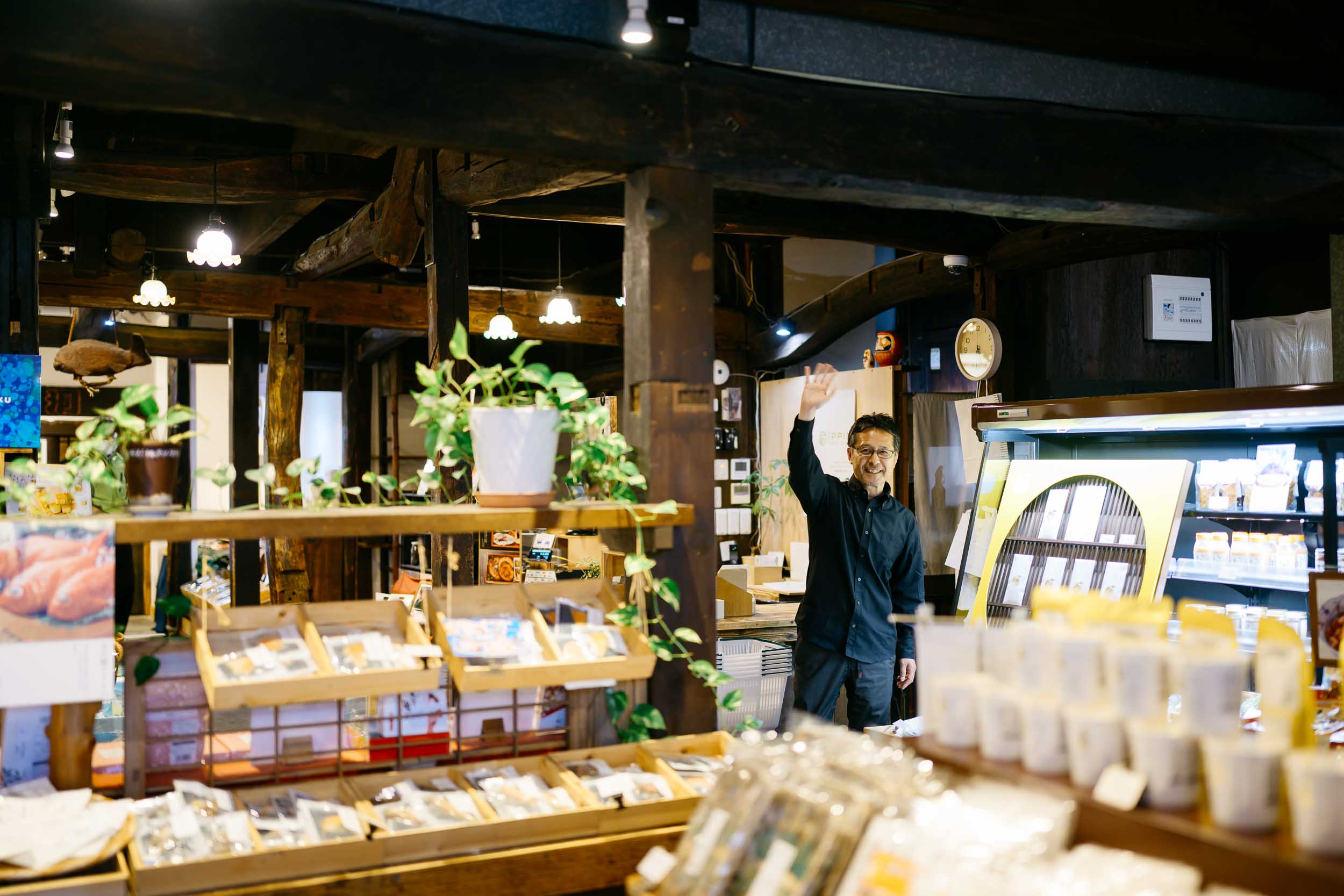 下田時計台フロントの木目調で温かみのある店内の様子。並べられたお土産ものの奥で、笑顔で手を振って出迎える代表の長池さんの写真。